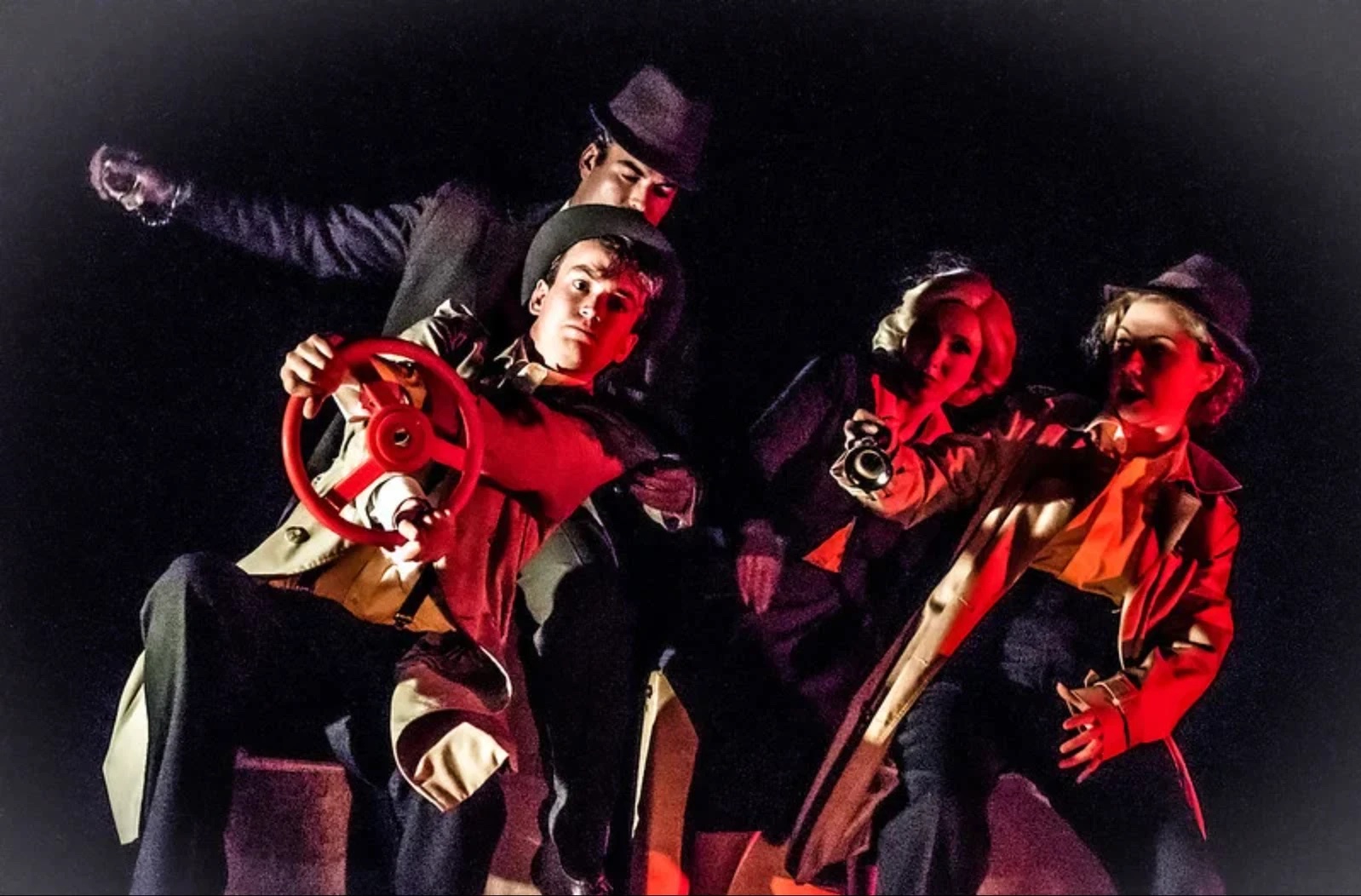 The car chase: four actors in hats and trench coats lit red, one gripping a steering wheel