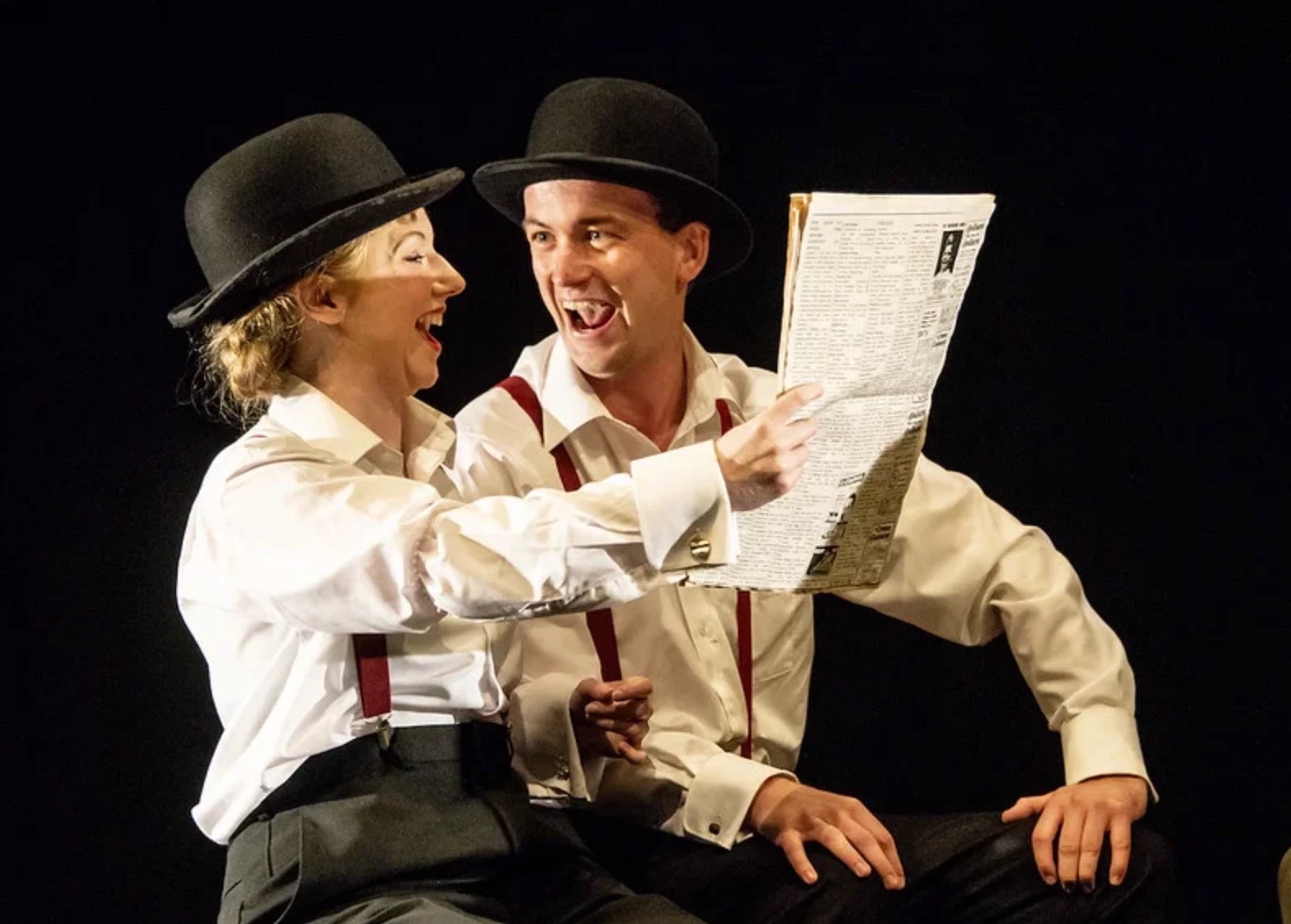 Two actors in bowler hats and braces laughing over a newspaper