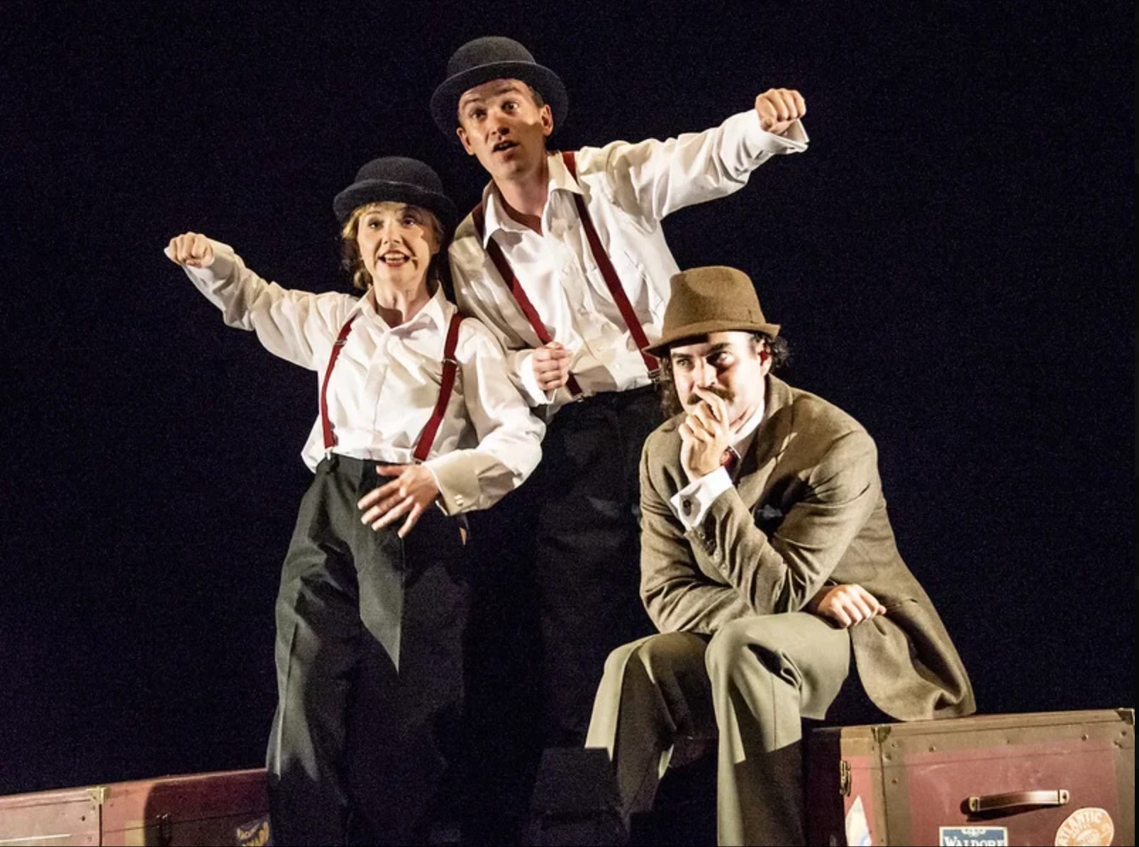 Two actors in bowler hats strike a pose behind Hannay, who sits on a trunk