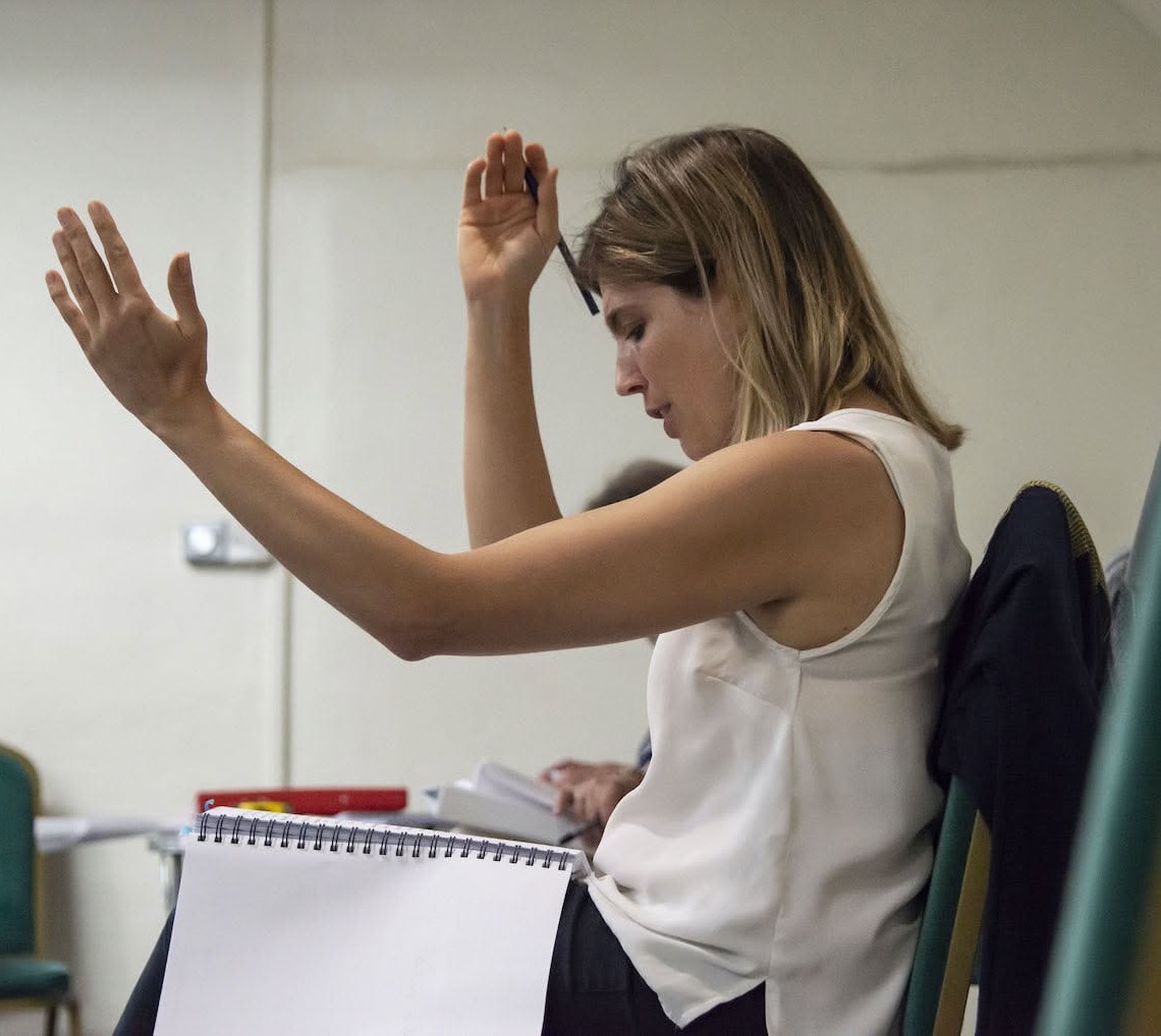 Estella Todisco in rehearsal, seated with a script on her lap, both hands raised